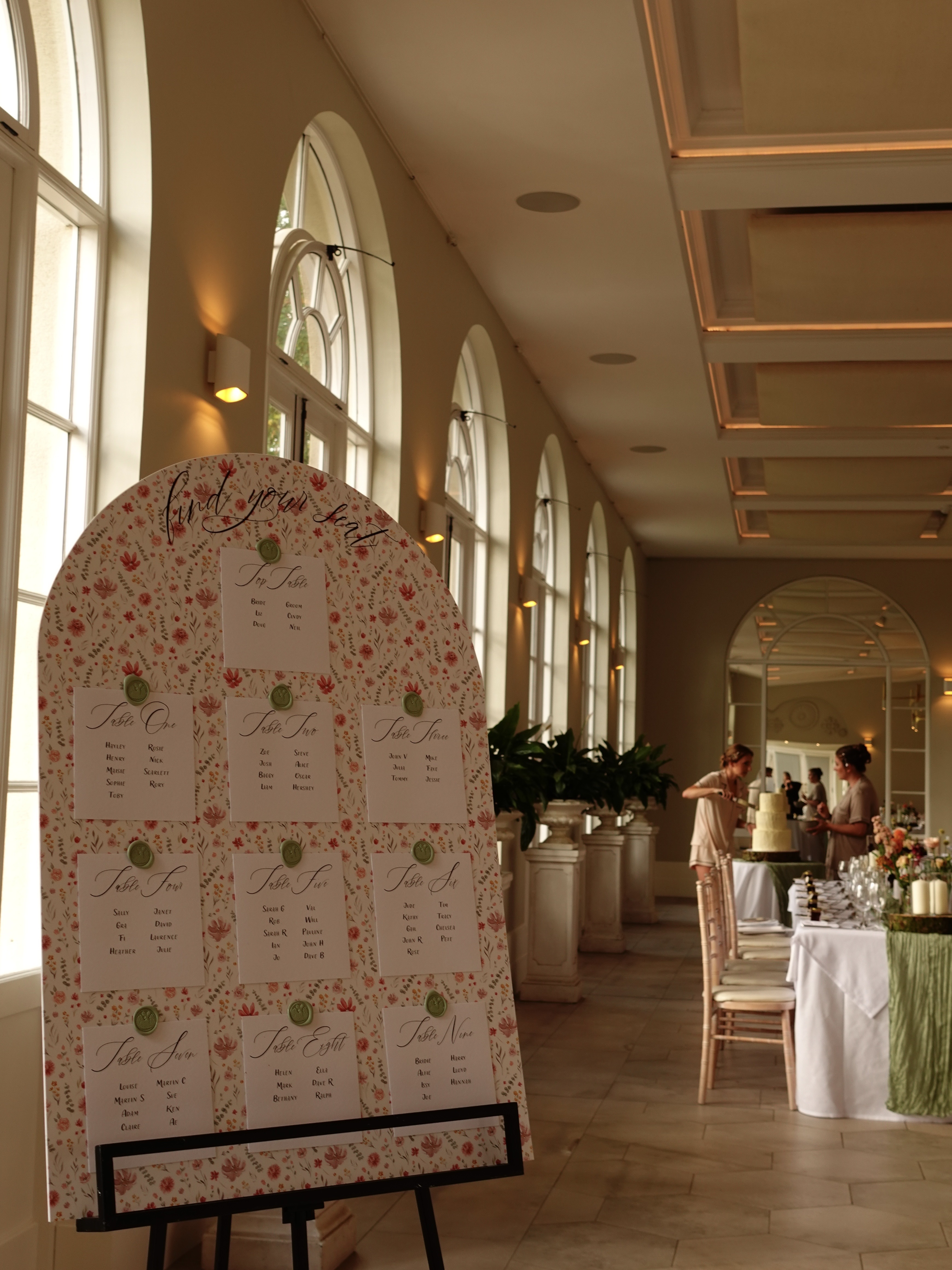 Floral wedding seating plan beside tall arched windows and reception tables